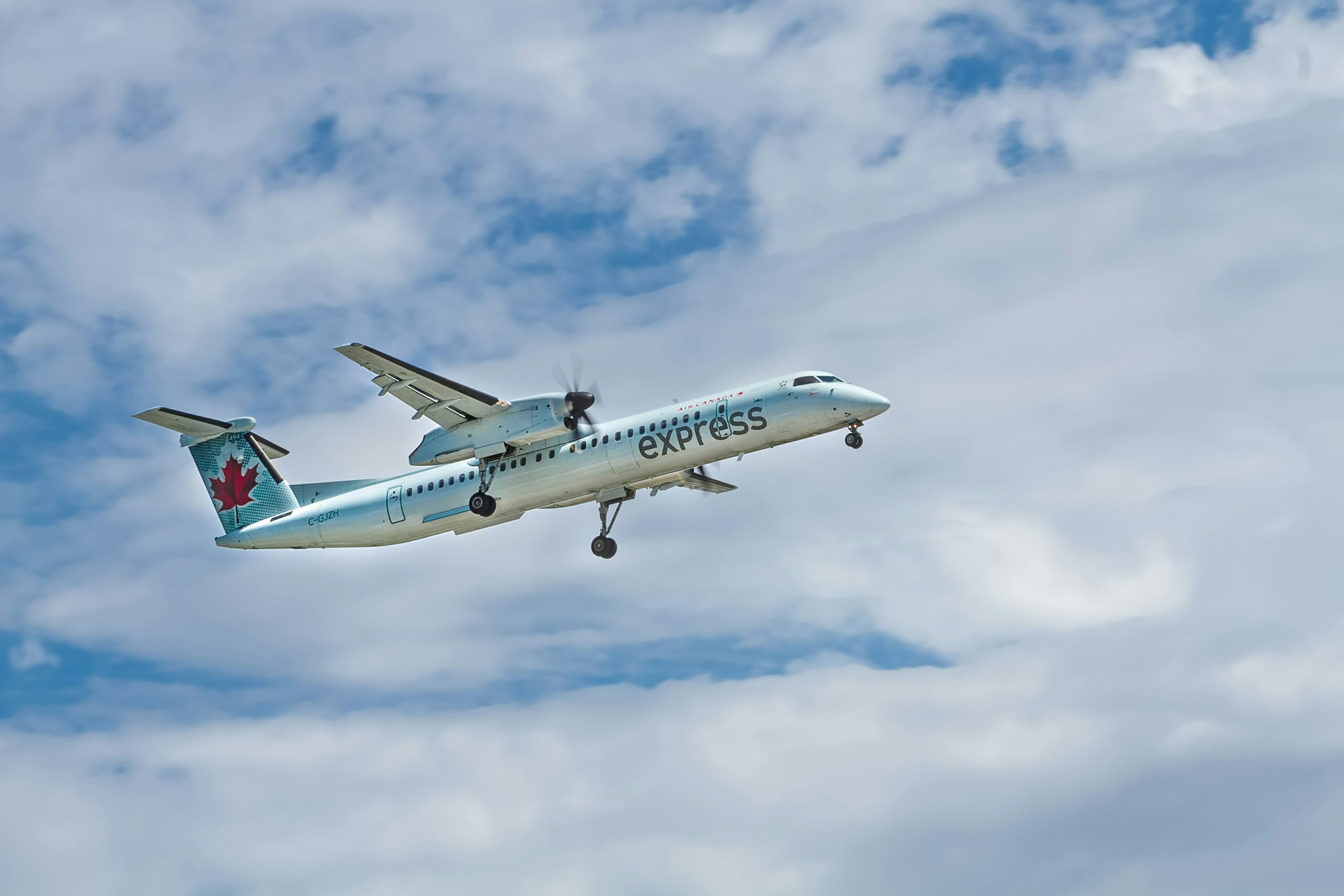 An Air Canada Express De Havilland Dash 8-400 climbs steadily through a cloudy blue sky. This modern turboprop aircraft is a staple of regional aviation, connecting smaller hubs with major international airports efficiently.